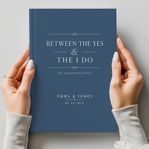 May include: A dark blue hardback engagement journal with white text that reads: BETWEEN THE YES & THE I DO, our engagement journal, EMMA & JAMES, and 05.24.2025. The book cover features thin horizontal lines and decorative flourishes.