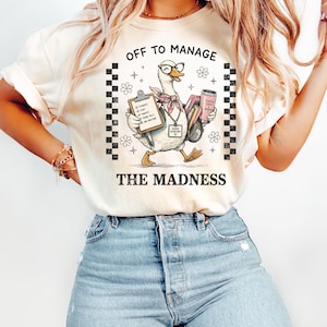 May include: Cream-colored t-shirt with the text "OFF TO MANAGE THE MADNESS." The graphic features a cartoon goose wearing glasses and a scarf, carrying a clipboard, books, and a coffee cup. Black and white checkered patterns are on the sides.