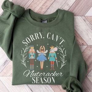 May include: A dark green sweatshirt with the text "SORRY. CAN'T. Nutcracker SEASON" in white. The graphic features a nutcracker, a ballerina, and a mouse king. The sweatshirt is folded, with the sleeves visible.