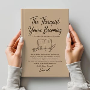May include: A tan hardcover journal titled The Therapist You're Becoming: A Journal for the Road to Licensure. The cover features a black line drawing of an open book, glasses, and a pen, with printed text below and the name Sarah at the bottom.