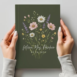 May include: A dark olive green hardcover journal featuring a central illustration of a wildflower bouquet with white daisies, pink cosmos, and purple lavender. The text "future Mrs. Parker" and "05.24.2025" is printed in white script and serif font.