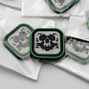 May include: A set of five green and white square patches with a black skull design. The patches have a white background with a black skull and a green border.