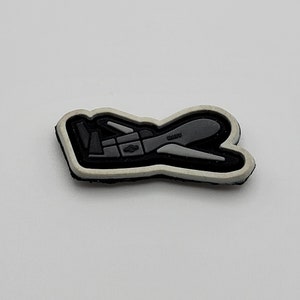 May include: A black and gray shoe charm with a stylized airplane design. The charm has a white border and is set against a white background. The word "USA" is visible on the airplane.