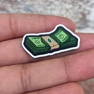 May include: A white and black rubber charm shaped like a stack of cash. The cash is green with a brown band and a letter C on the top bill.