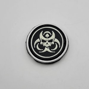 May include: A round, black patch with a white skull and biohazard symbol design. The patch has a white border and is likely made of rubber or a similar material. The design is centered on the patch.