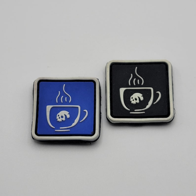 K&ouml;nnte beinhalten: Zwei quadratische Gummi-Charms. Einer ist blau mit einer wei&szlig;en Umrandung einer Kaffeetasse mit einem Totenkopf im Inneren und aufsteigendem Dampf. Der andere ist schwarz mit dem gleichen Design. Beide haben einen wei&szlig;en Rand.