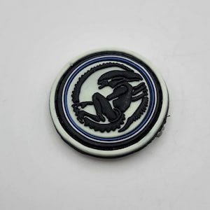 May include: A black and white alien creature graphic on a white round patch with blue and white concentric circles.