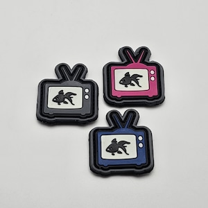 May include: Three iron-on patches in the shape of a retro television set with a black silhouette of a goldfish inside. The patches are in gray, pink, and blue.