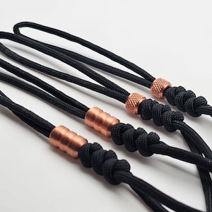Könnte beinhalten: Drei schwarze Paracord-Lanyards mit Kupferperlen. Die Perlen sind in verschiedenen Größen und Formen. Die Lanyards sind an den Enden verknotet.