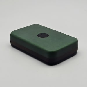 Puede incluir: Un objeto rectangular de color verde oscuro con bordes redondeados y un pequeño detalle circular negro en la superficie superior. El borde inferior es de color marrón oscuro. El objeto es probablemente un pequeño dispositivo o accesorio electrónico.