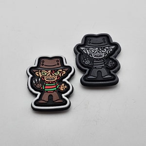 Może przedstawiać: Dwa czarno-białe łatki do prasowania z postacią z kreskówki z nożem. Postać po lewej stronie ma na sobie czerwoną i zieloną paskowaną koszulę i brązowy kapelusz. Postać po prawej stronie ma na sobie czarno-białą paskowaną koszulę i czarny kapelusz.