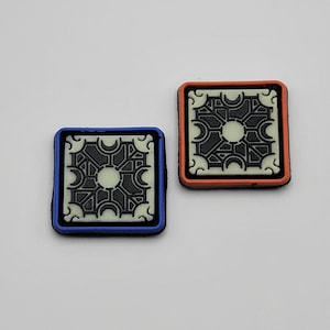 May include: Two square patches with a black and white geometric design. The patch on the left has a blue border and the patch on the right has an orange border.