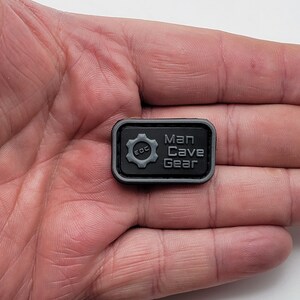 Pu&ograve; includere: Toppa rettangolare nera con un ingranaggio grigio e il testo "Man Cave Gear" e "EDC" all'interno dell'ingranaggio.