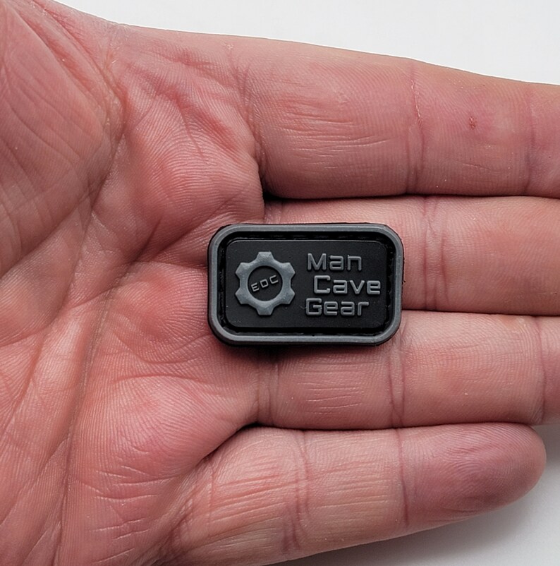 Pu&ograve; includere: Toppa rettangolare nera con un ingranaggio grigio e il testo "Man Cave Gear" e "EDC" all'interno dell'ingranaggio.