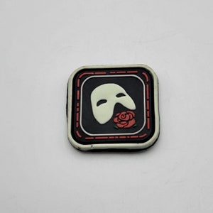 Op de afbeelding: Een zwart-witte patch met een wit spook van de opera-masker en een rode roos. De patch is omgeven door een rode en witte rand.