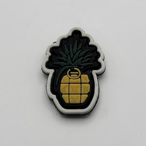 Può includere: Una toppa bianca e nera con un design di ananas verde. L'ananas è una granata con una miccia gialla e uno spillo nero.