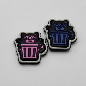 Pode incluir: Dois pequenos patches de borracha preta com contorno branco. Cada patch apresenta um guaxinim de desenho animado segurando uma lata de lixo. Um guaxinim é rosa e o outro é azul.