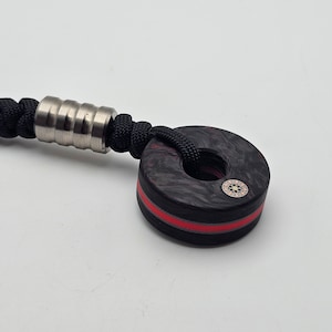 Könnte beinhalten: Ein schwarzer und roter Ring mit einem kupfernen Mittelstück. Der Ring ist an einer schwarzen Schnur mit einer silbernen Perle befestigt.