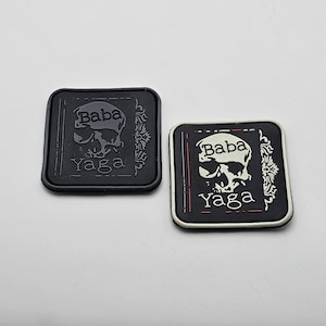 Puede incluir: Dos parches bordados en blanco y negro con un diseño de calavera y huesos cruzados. Los parches presentan el texto "Baba Yaga" en letras blancas.