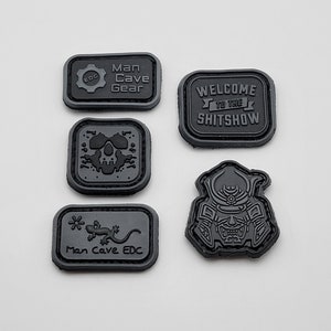 May include: Five black and gray embroidered patches with different designs. The patches include a skull, a lizard, a samurai, a gear with the text "Man Cave Gear", and a banner with the text "Welcome to the Shitshow".