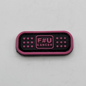 Pode incluir: Remendo de tecido rosa e preto em forma de bandagem com o texto "F#U CANCER" em branco.