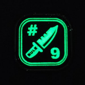 May include: A green glow-in-the-dark patch with a square shape and a black background. The patch features a stylized knife with the word "RULE" on the handle, a number "9", and a hashtag symbol.