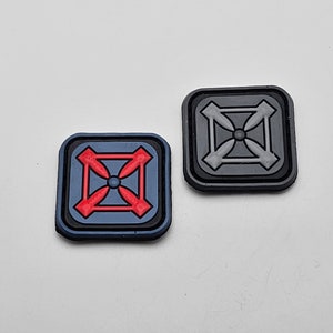 Könnte beinhalten: Zwei quadratische Patches mit einem Kreuzdesign. Ein Patch ist grau mit einem weißen Kreuz, und der andere ist blau mit einem roten Kreuz.