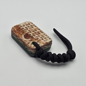 May include: A rectangular, wooden fidget toy with a woven pattern on top. The toy has a green stripe around the middle and is attached to a black cord. The wood has a natural, light brown color.