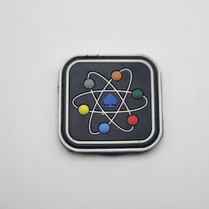 May include: Square patch with a black background and a white border. The patch features a colourful atom design with a blue spade symbol in the centre. The atom has a white orbital design with colourful spheres.