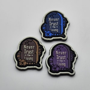 May include: Three tombstone-shaped patches with the text "Never Trust ~The~ Living" in white lettering. The patches are in blue, brown, and purple.