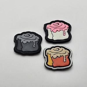 May include: Three different colored felt patches shaped like cinnamon rolls. The patches are gray, white, and orange. Each patch has a swirl design on top.
