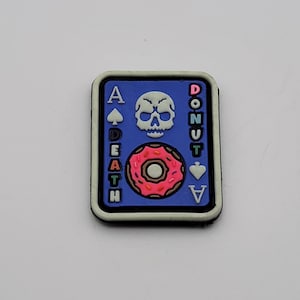 May include: A blue, rectangular charm with a black border. The charm features a skull, a pink donut, and the words "A DEATH" and "DONUT A" in white. The charm is a novelty item.
