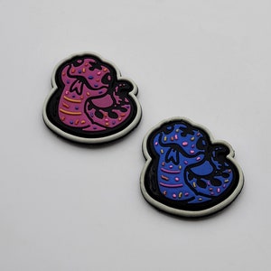 May include: Two colorful shoe charms. One is pink with purple sprinkles and the other is blue with blue sprinkles. Both charms have a black border and a white outline. The charms are shaped like a stylized snake.