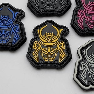 Puede incluir: Primer plano de varios parches negros con un diseño de casco de samurái. Los parches presentan varios esquemas de color, incluyendo azul, amarillo, rosa y blanco, con contornos negros. Los parches son probablemente para ropa o accesorios.