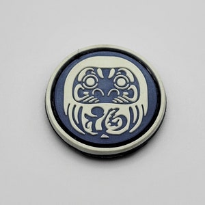 May include: A round, white, plastic patch with a dark blue outline. The patch features a white, stylized image of a Japanese Daruma doll with a black outline.