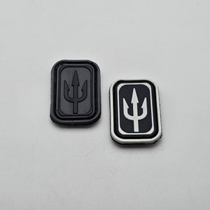 Może przedstawiać: Dwa prostokątne czarno-białe zawieszki do butów. Jeden jest czarny z czarnym symbolem trójzębu. Drugi jest czarny z białym symbolem trójzębu i białym obramowaniem. Oba zawieszki mają wypukły wzór.