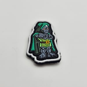 Op de afbeelding: Een zwart-wit cartoon patch van Doctor Doom, een superschurk uit Marvel Comics. Hij draagt een groene cape en een gele riem. De patch is gemaakt van rubber en heeft een witte omtrek.