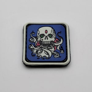 Puede incluir: Parche cuadrado con un diseño de calavera y tentáculos sobre fondo azul. La calavera tiene ojos verdes y detalles rosas. El parche tiene un borde blanco.