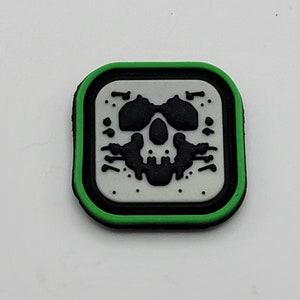 May include: A black and white skull graphic on a white background with a green border. The patch is made of rubber.