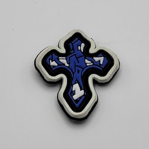 May include: A blue and white cross with a white outline. The cross is made of a rubber-like material and has a textured surface.