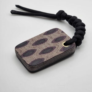 Op de afbeelding: Een kleine, rechthoekige sleutelhanger met een zwart en bruin luipaardprintontwerp. De sleutelhanger heeft een zwart koord met een knoop aan de bovenkant en een gouden metalen ring.