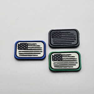 Op de afbeelding: Drie kleine Amerikaanse vlagpatches, een met een blauwe rand, een met een groene rand en een met een zwarte rand.