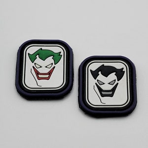 May include: Two square patches with a white background and a black outline. Each patch features a cartoon Joker face. One Joker has green hair and a red smile. The other Joker has black hair and a white smile.