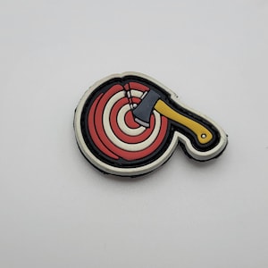 May include: A shoe charm featuring a target with red and white concentric circles, a black border, and a yellow-handled axe. The axe is gray and black, and the charm has a white background.