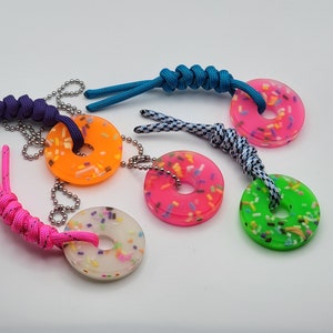 May include: Five colorful donut-shaped keychains with sprinkles. Each keychain is attached to a string or cord. The keychains are pink, orange, white, green, and blue.