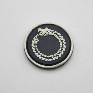May include: A black and white circular patch with a white dragon design. The dragon is curled in a circle, forming a ring. The patch has a white border.