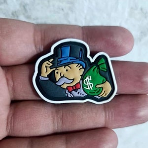 Puede incluir: Una figura de Monopoly Man en blanco, negro, azul, verde y rojo sosteniendo una bolsa de dinero verde con un signo de dólar.
