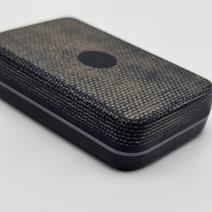 Puede incluir: Un estuche rectangular de color oscuro con un patrón texturizado similar al tejido en la superficie superior. El estuche tiene una pequeña hendidura circular y una fina franja de color claro a lo largo de los bordes. El diseño es simple y moderno.