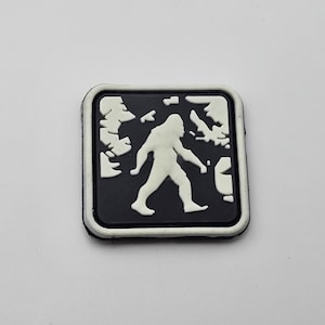 Könnte beinhalten: Schwarz-weiß-Patch mit einer weißen Silhouette von Bigfoot, der durch einen Wald geht. Der Patch hat einen weißen Rand.