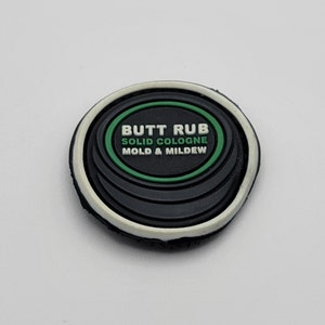 以下が含まれることがあります： 「BUTT RUB SOLID COLOGNE MOLD & MILDEW」のテキストが入った、黒、白、緑の丸い固形香水。デザインは層状になっています。